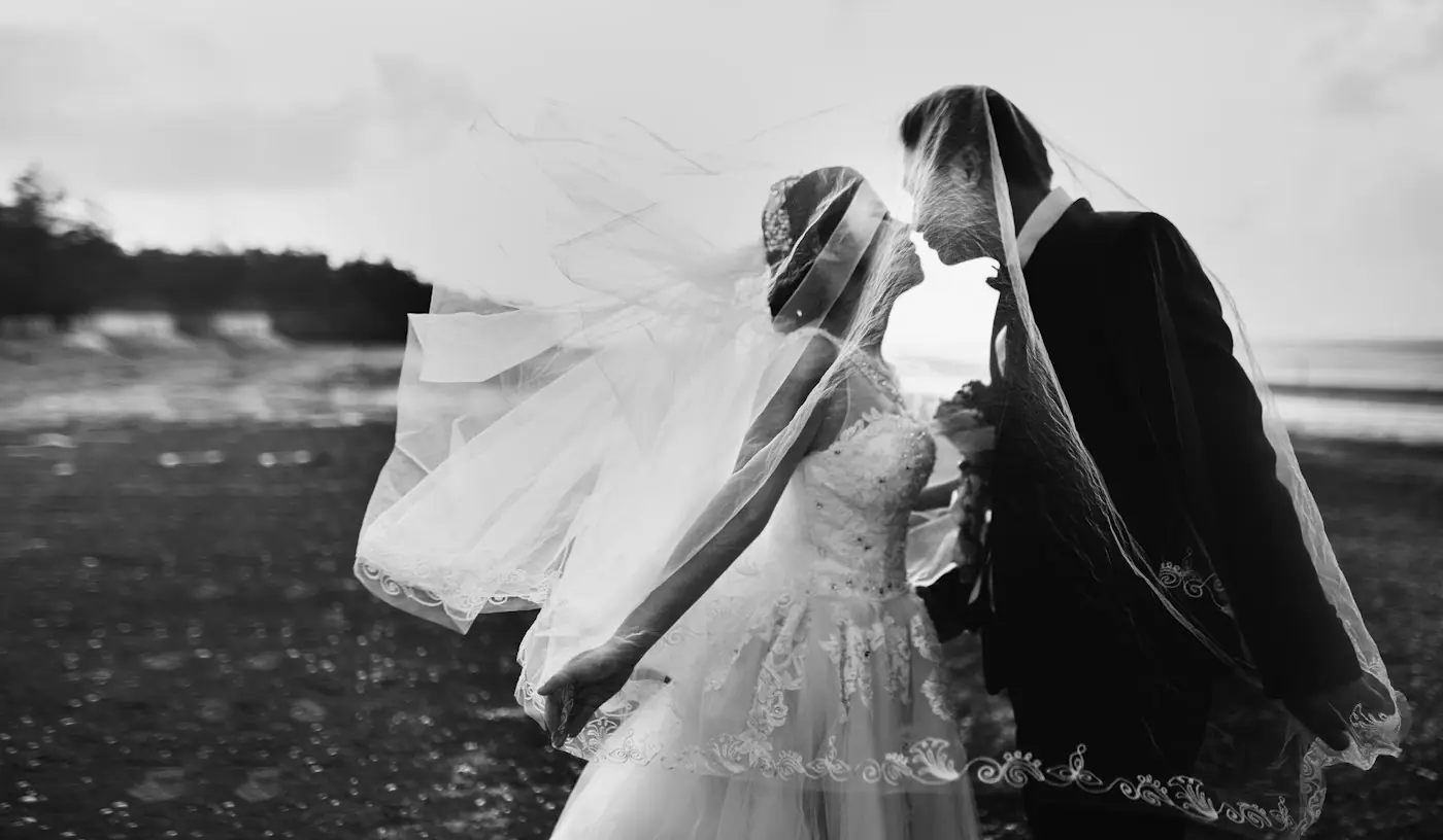 Black and white photo of a bride and groom kissing as her veil lifts in the wind